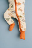 A close-up of a baby footed one-piece sleeper with an orange zipper, cream background, and tractor pattern, shown against a solid blue-gray background.