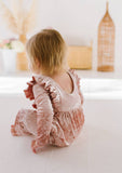 Child wearing a pink velvet ruffled romper with scoop back sitting on a light-colored floor.