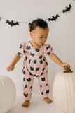 Child in pink outfit with black cat pattern holding a white pumpkin, surrounded by Halloween decorations.