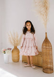 Young girl in a pink velvet dress standing between two tall wicker vases with dried plants against a white wall.