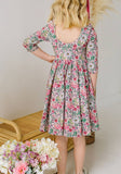 girl wearing a long sleeve floral dress with a scoop back standing backward with a white background and flowers in the foreground