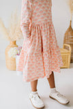 A child wearing a tiered long-sleeve midi dress with a pink floral pattern and pockets, paired with white shoes, standing in a brightly lit room with decorative dried grass and wicker baskets in the background.