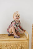 Baby in a floral long sleeve bubble romper sitting on a wicker stool with a white background