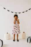 Child in a pink dress with black cat pattern standing in front of Halloween decorations including pumpkins and a bat banner.