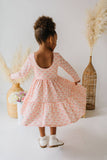 A young child with a high bun hairstyle is shown from behind wearing a light pink tiered dress with a geometric pattern. The child is standing in a bright room with decorative dried pampas grass in woven baskets.