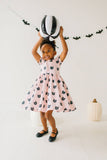 Child in a pink dress with black cat patterns holding a black and white striped balloon against a white background.