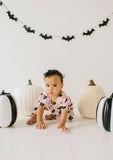 Baby in a pink outfit with black cat patterns surrounded by Halloween-themed decor including pumpkins and bat decorations.