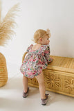 Child in a long sleeve floral bubble romper with scoop back  standing next to a wicker bench with pampas grass in the background