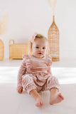 Baby in a long sleeve pink velvet romper sitting on a white surface with wicker baskets in the background.