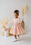 A young girl with a high bun wearing a pink and white patterned long-sleeve dress, white shoes, and a hair bow, standing in a bright studio with pampas grass and woven baskets.