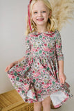 Young girl wearing a floral dress with long sleeves and  a peter pan collar, standing against a neutral background.