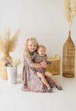 Two young girls in matching floral romper and  dress sitting together with decorative elements like a woven basket and pampas grass.