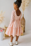 A young girl with her hair in a high bun is shown from behind, wearing a light pink long-sleeve tiered dress with a floral pattern. She is standing in a styled indoor setting with a wicker basket, dried pampas grass, and a white vase of flowers.