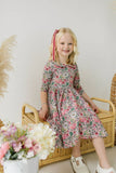 Young girl in a floral dress with long sleeves and a peter pan collar sitting on a wicker chair with flowers and a plant in the foreground.