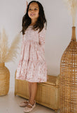 Young girl in a pink velvet dress standing in front of wicker furniture and decorative plants.