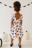 Child wearing a pink dress with black cat patterns in a Halloween-themed setting.