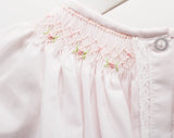 Close-up of a light pink baby garment with delicate floral embroidery, a smocked neckline, and white lace trim.
