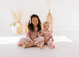 Two children in matching pink velvet outfits sitting on a white surface with decorative elements in the background.