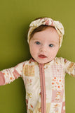 A baby wearing a cream and pink patchwork zip-up onesie and a matching patterned headband, standing against a solid olive green background.