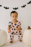 Baby wearing a pink romper with black cat prints, standing between two white pumpkins with a Halloween bat banner in the background.