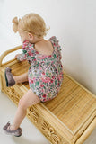 Child in a floral bubble romper sitting on a wooden step stool against a white wall.