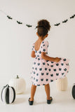 Child wearing a pink dress with black cat patterns, standing in front of Halloween decorations.