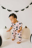 Child in pink Halloween-themed onesie with pumpkins and bats, standing in front of white pumpkins and a bat banner.