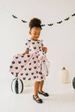 Child wearing a pink dress with black cat patterns, standing in a room decorated for Halloween.