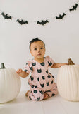 Baby in a pink romper with black cat patterns sitting between two white pumpkins, with Halloween decorations in the background.