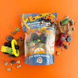 Monster Truck KidDough Play Kit in a clear plastic bag with blue and orange graphics, surrounded by toy vehicles, small rocks, and dirt mounds on an orange background.
