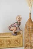 Child in floral bubble romper standing on a wicker bench with a textured wall and vase in the background
