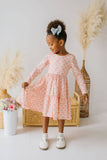 A young girl with her hair in a high bun wearing a light pink long-sleeve tiered dress with a geometric pattern. She is standing in a studio setting with decorative pampas grass, a wicker chest, and a white flower pot.