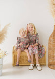 Two young girls in matching floral outfits sitting on a wicker stool with a white background.