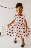 Young girl wearing a pink dress with black cat patterns, standing in a room with Halloween decorations.