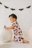 Baby in pink romper with black cat pattern playing with a white pumpkin against a white background with bat decorations.