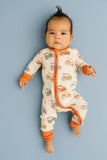 A baby lying on their back wearing a cream-colored footless zip-up sleeper with orange cuffs and a tractor pattern, set against a solid blue background.