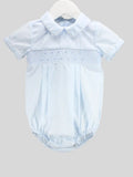 A light blue baby romper with a short-sleeve collar, featuring a smocked chest panel and a pleated bodice, hanging on a white hanger against a solid gray background.