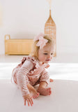 Young child in a long sleeve pink velvet romper with shoulder ruffles sitting on a white surface.