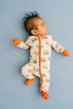 A baby lying on their back wearing a footed zip-up sleeper with an orange zipper, orange mittens, and an orange tractor pattern on a cream background, set against a solid light blue background.